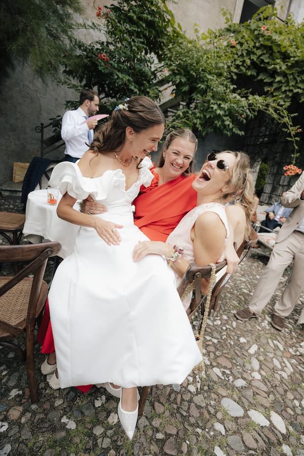 Bride in white dress laughing with two female guests in red and white outfits at an outdoor wedding reception.