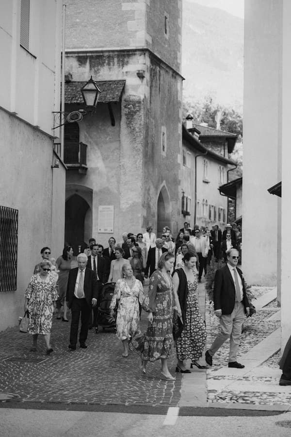 Black and white photo of elegantly dressed wedding guests walking through a charming Italian village street.