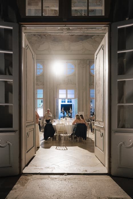 Elegant dinner party viewed through grand white double doors in an ornate classical venue with glowing candlelight.