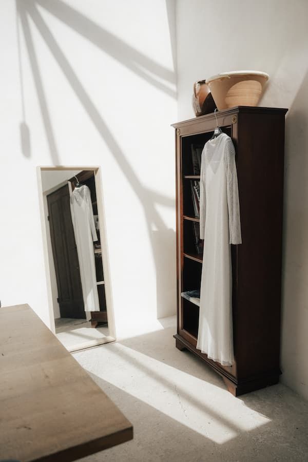 White dress hanging in dark wood armoire beside floor mirror with sunlight shadows on white walls.