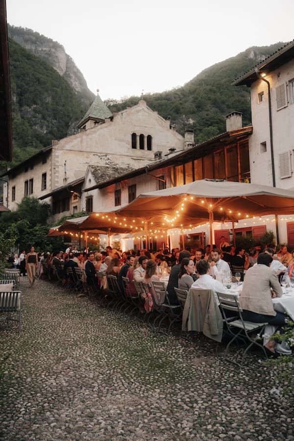 Outdoor wedding reception dinner along cobblestone courtyard with string lights, long tables, and mountain backdrop at dusk.