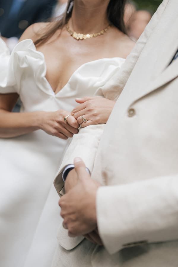 Bride in white gown with gold necklace and rings holds groom's arm in beige suit at wedding ceremony.
