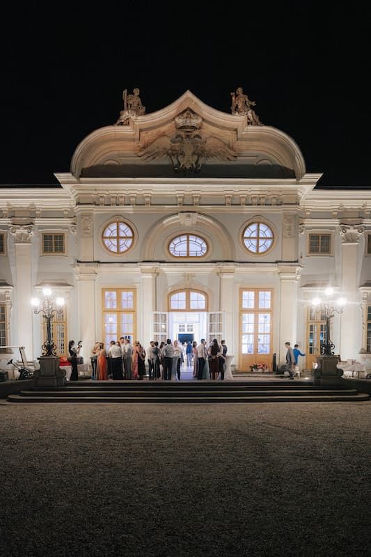 Elegant white baroque palace illuminated at night with guests gathered on steps for a formal evening event.