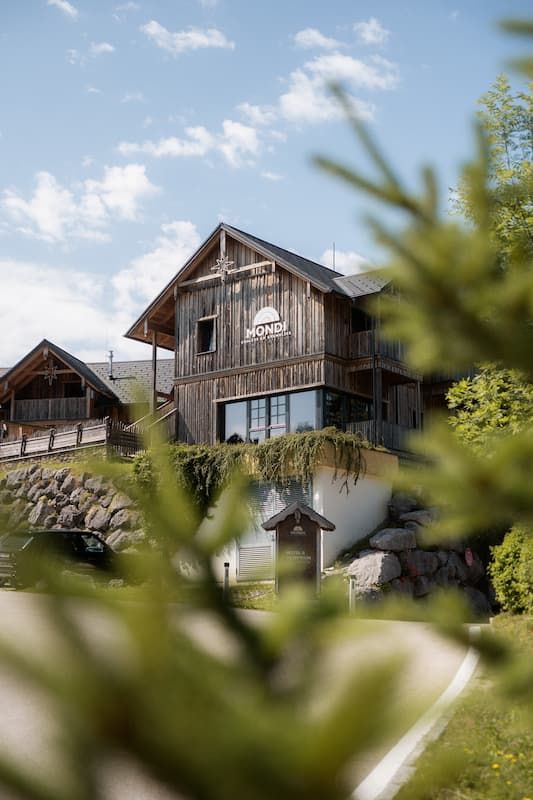 Rustic wooden Mondi hotel building surrounded by lush greenery and stone walls under a blue sky.