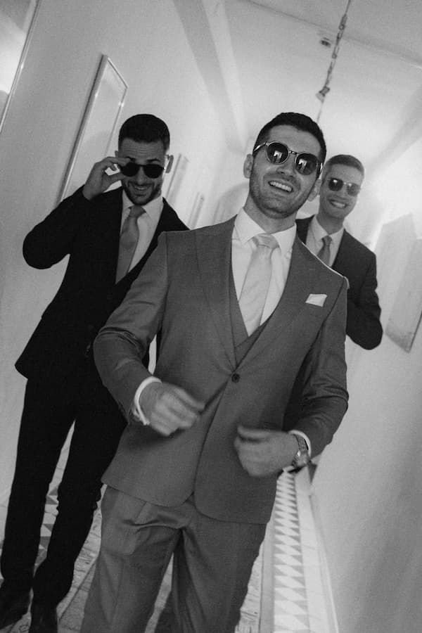 Three stylish groomsmen in suits and sunglasses pose confidently in a hallway in this black and white wedding photo.