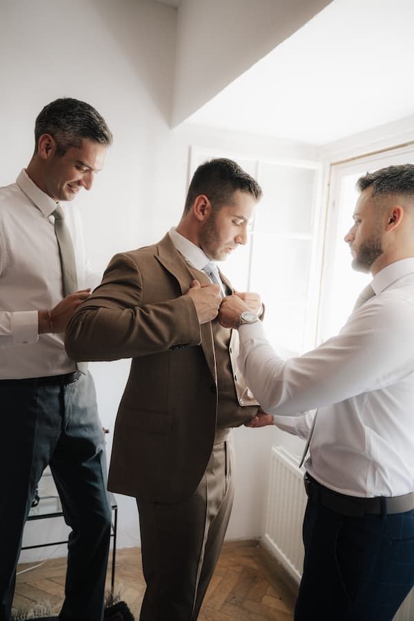 Groom in brown suit being helped by two groomsmen in white shirts adjusting his jacket before wedding ceremony.