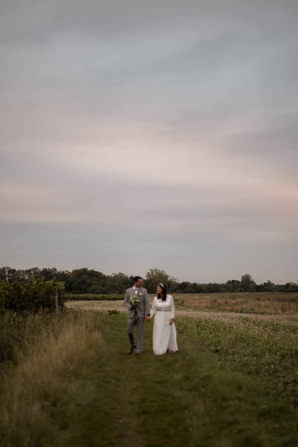 Elena & Michael gehen Hand in Hand über eine Wiese. Der Himmel ist voller rosafarbener Wolken.
