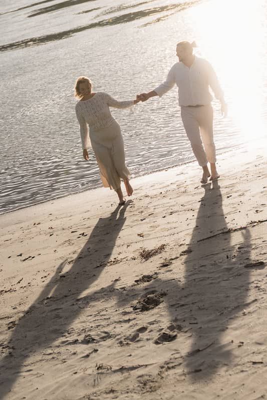 Doni & Babsi gehen Hand in Hand über den Strand. Die Sonne hinter ihnen lässt lange Schatten über den Sand fallen