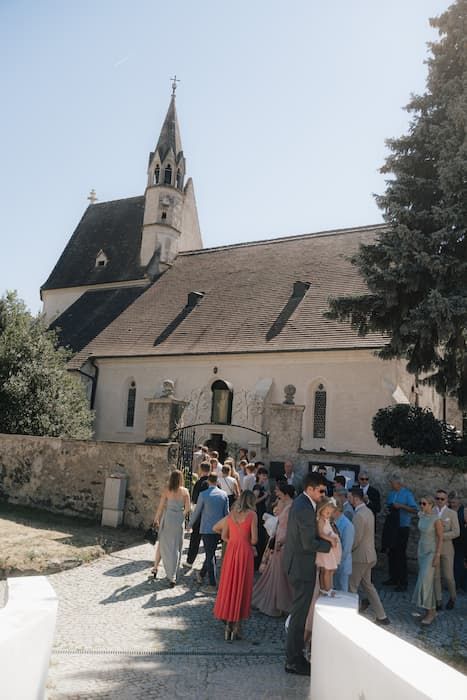 Guests gather outside a historic stone church with a Gothic spire on a sunny day for a wedding ceremony.
