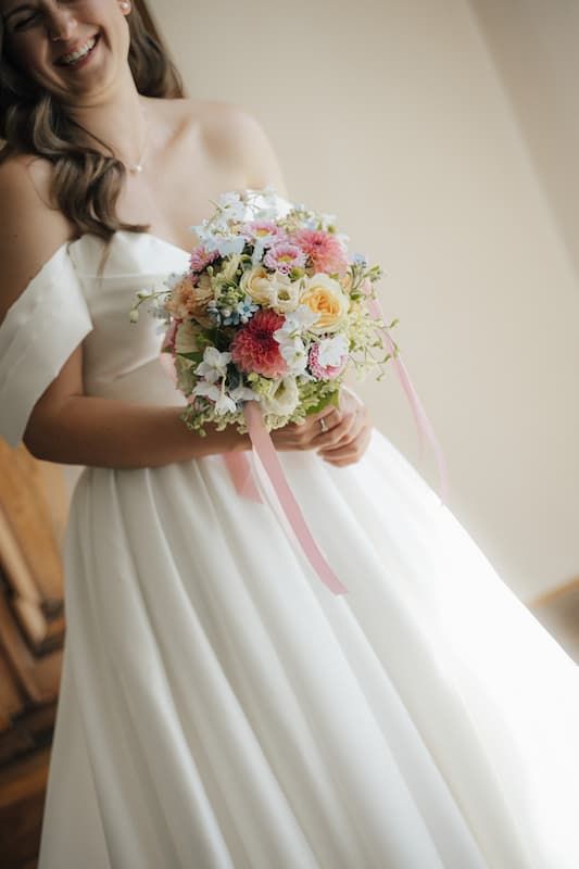 Smiling bride in white off-shoulder gown holding colorful wedding bouquet with pink ribbons and mixed flowers.