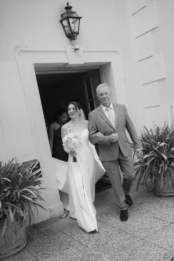 Black and white photo of a bride in a white gown holding a bouquet, walking with an older man in a suit from a building.