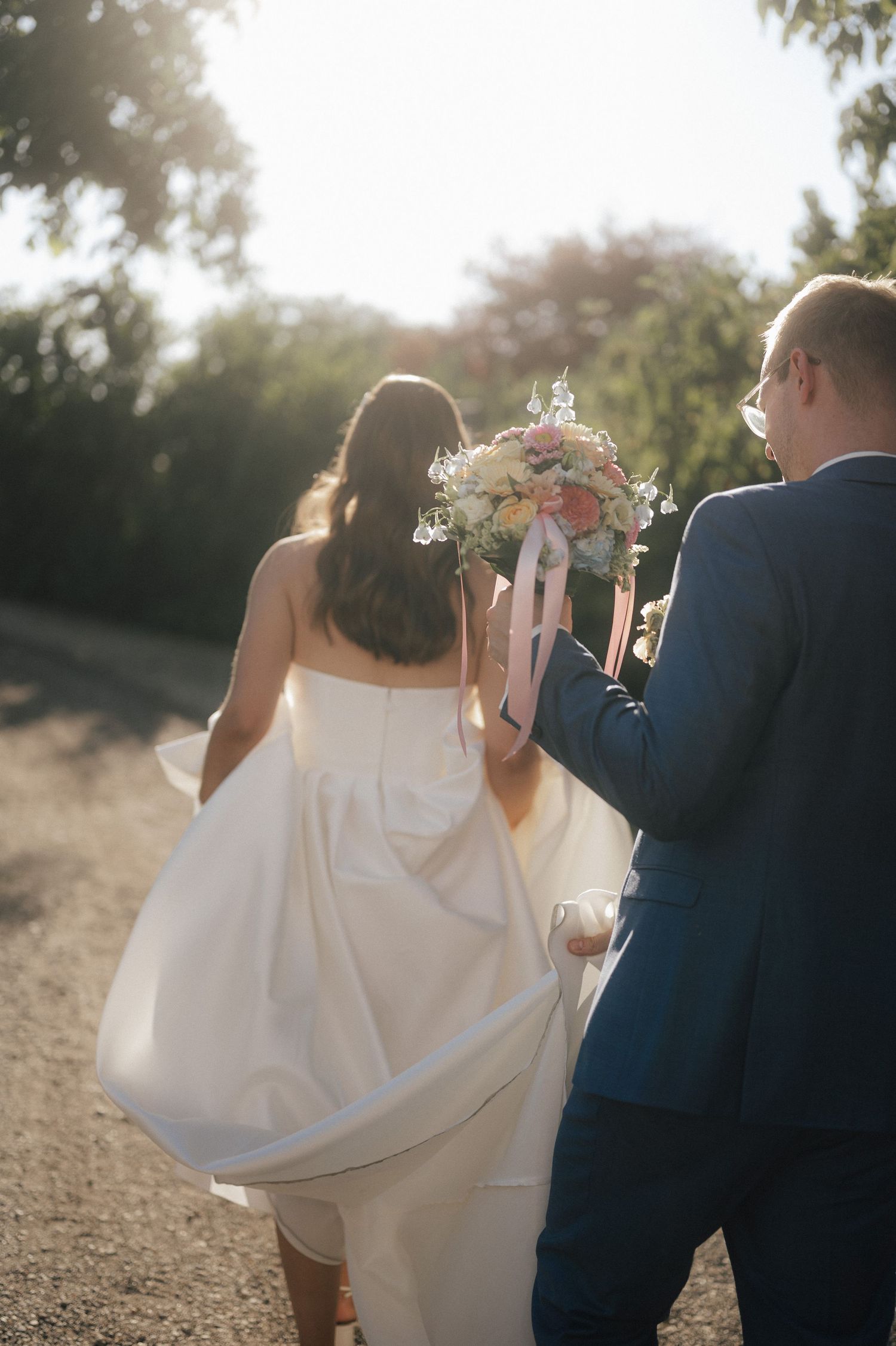 Bride and groom walking away on sunny path, bride in white dress holding pastel bouquet, groom in blue suit.