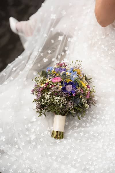 Ein Wildblumen-Brautstrauß mit bunten Blumen liegt auf dem Hochzeitskleid neben der Braut.