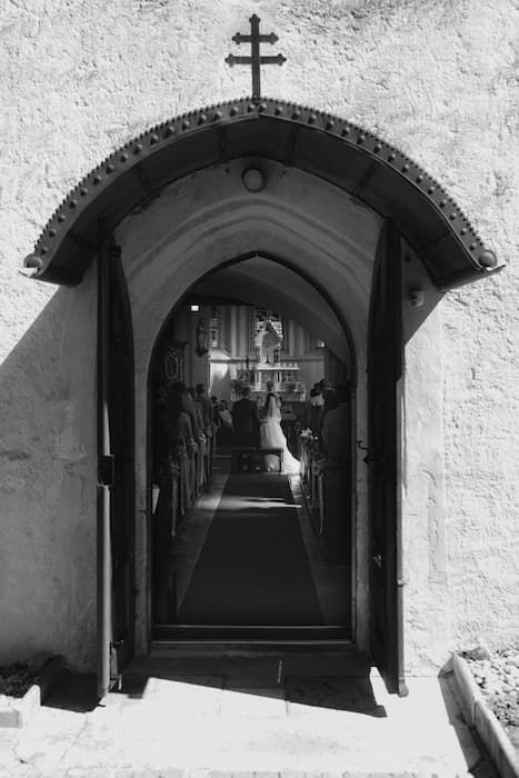 Black and white photo of a church doorway with cross, revealing a wedding ceremony inside with bride at the altar.