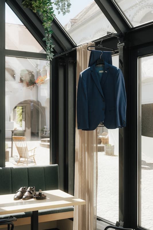 Blue suit jacket hanging in a sunlit glass conservatory restaurant with leather seating and dress shoes on a wooden table.
