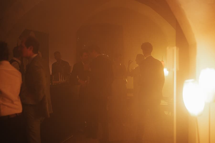 Atmospheric party scene with warm golden light and fog, guests socializing in a dimly lit venue.