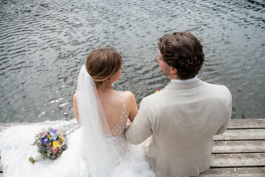 Bride and groom sitting on wooden dock by lake, bride holding colorful bouquet, wearing white gown and veil.