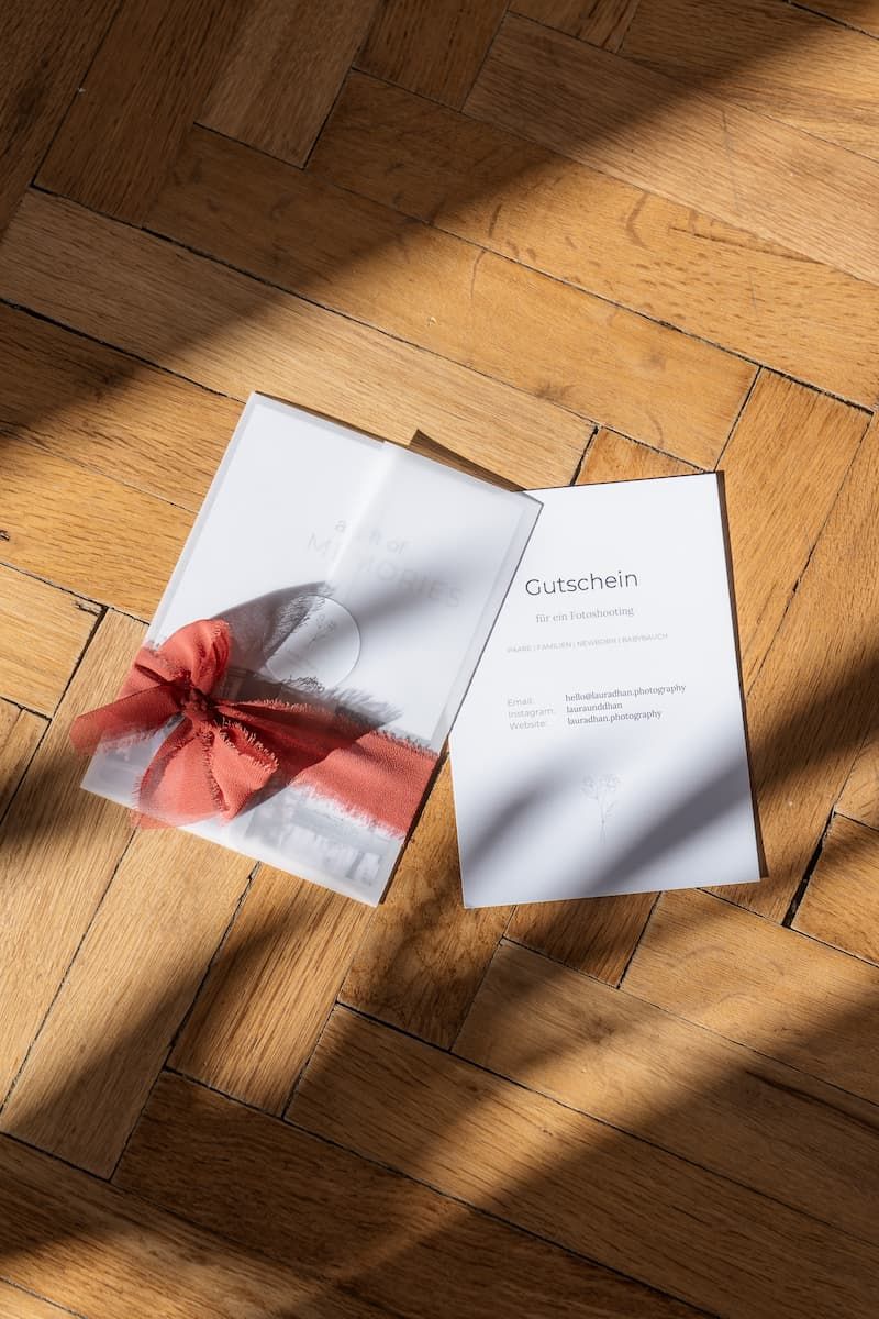 Wedding photography gift voucher with red bow and card displayed on a sunlit herringbone wood floor.