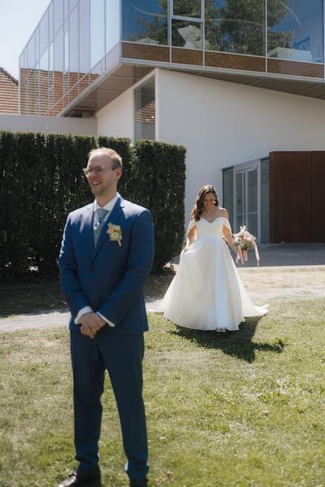 Groom in blue suit stands on lawn as bride in white ball gown approaches during outdoor first look wedding moment.