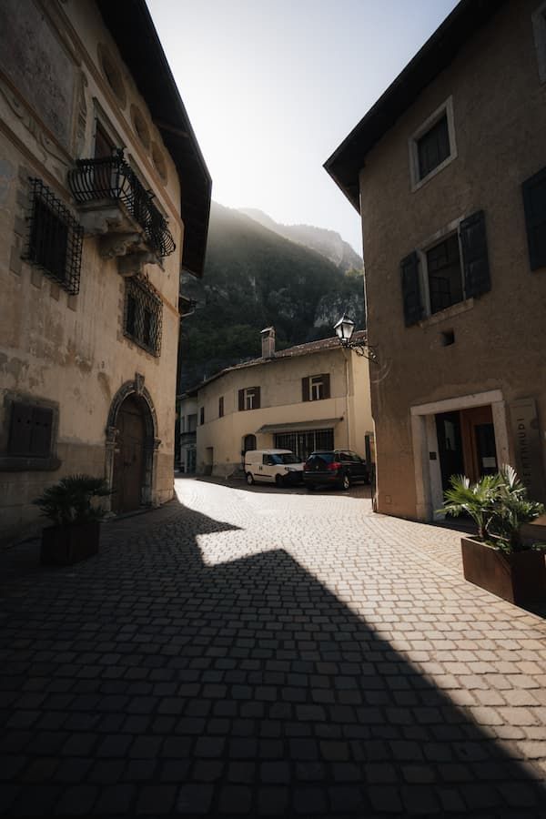 Cobblestone street in a historic Alpine village with old stone buildings, parked cars, and misty mountains in the background.