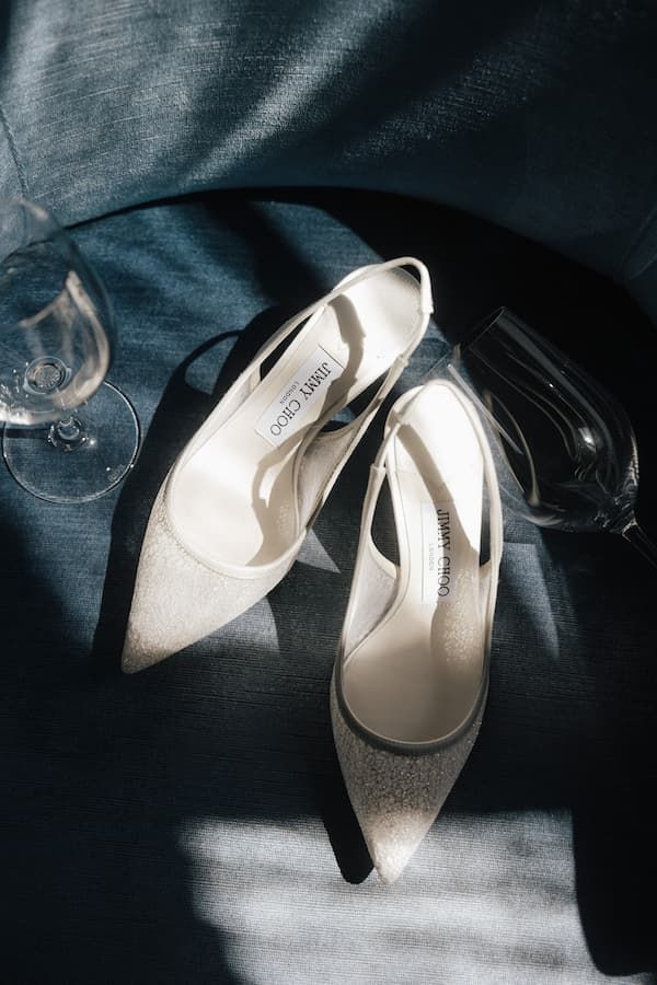 Elegant white Jimmy Choo slingback heels displayed on blue fabric beside crystal champagne glasses in soft sunlight.