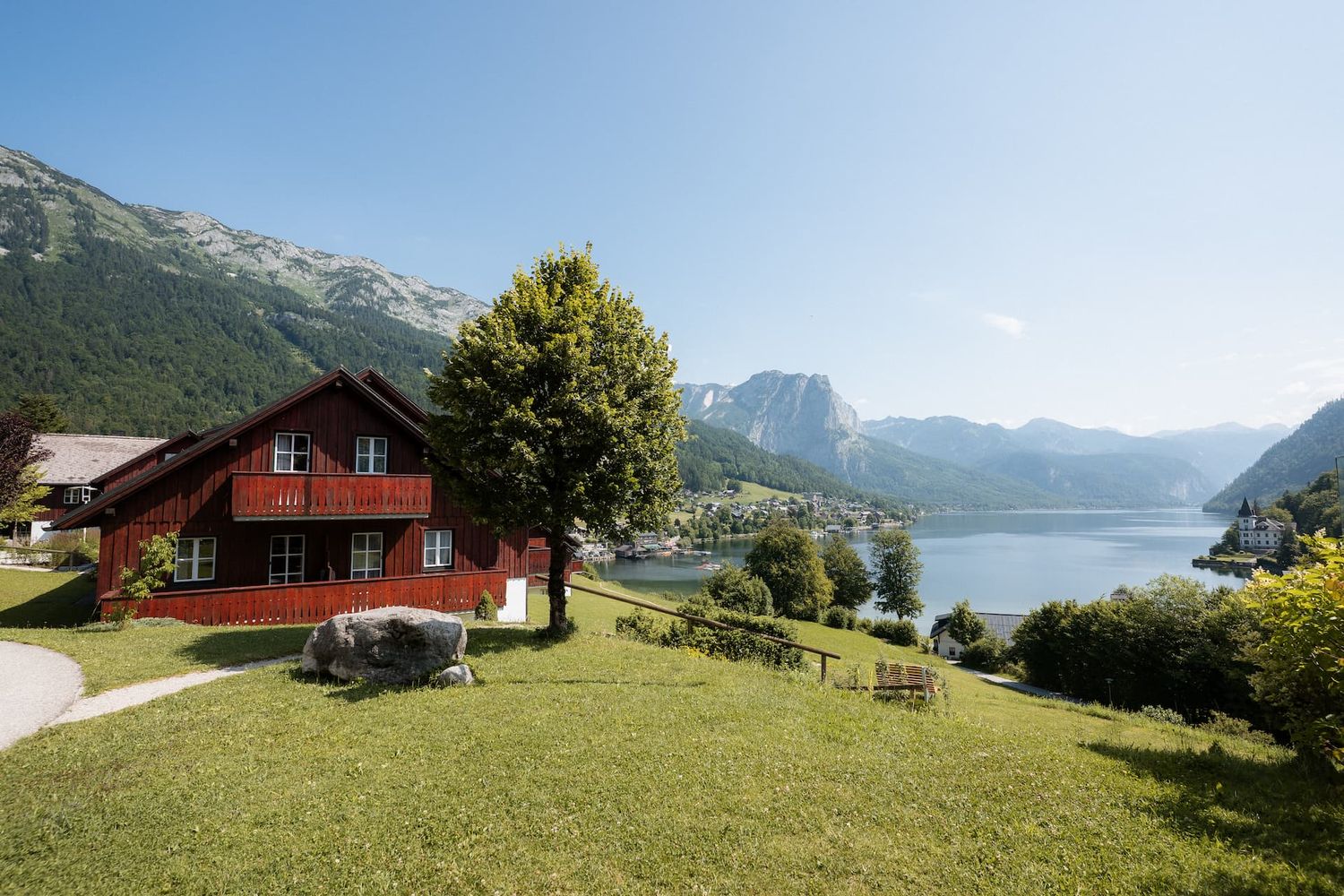 Red alpine chalet with mountain lake views, lush green lawn, and scenic Austrian Alps backdrop on a sunny day.