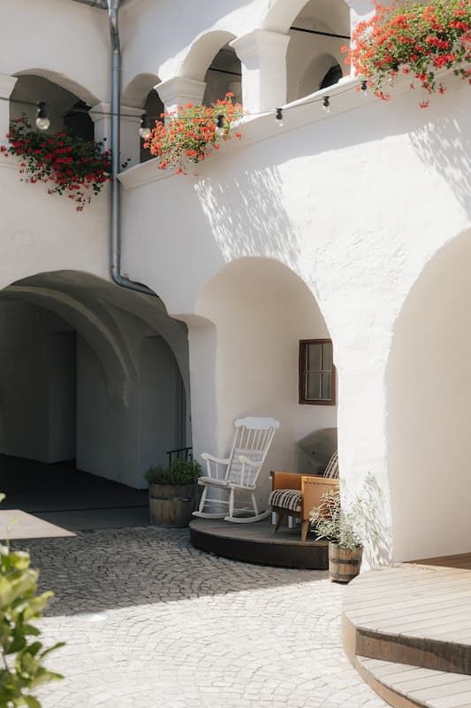 White Mediterranean courtyard with arched walls, red geraniums, rocking chair, and cobblestone paving in sunny setting.