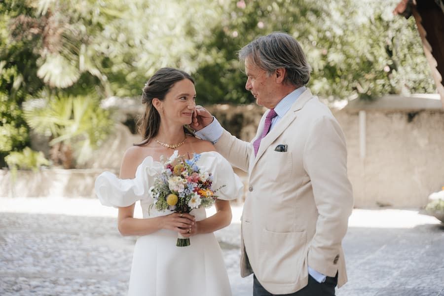 Bride in white off-shoulder gown holds colorful bouquet as older man in beige suit gently touches her face outdoors.
