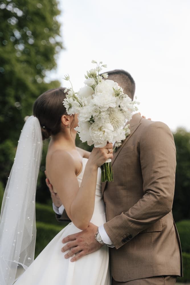 Bride and groom kissing behind white peony bouquet outdoors, bride in backless gown with veil, groom in tan suit.