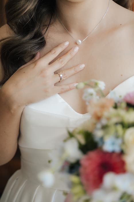 Bride in white strapless gown displays gold wedding ring, holding colorful bouquet with pearl necklace.