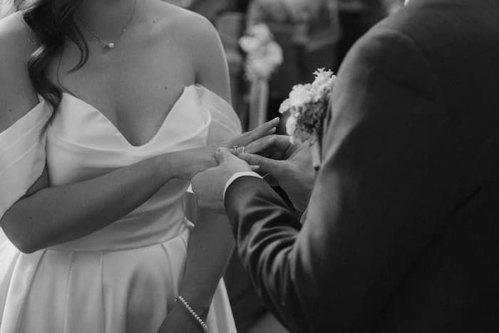 Black and white photo of a groom placing a wedding ring on the bride's finger during a wedding ceremony.
