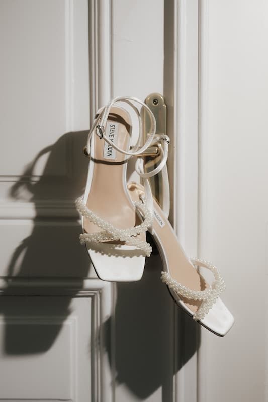White pearl-embellished block heel sandals with ankle strap displayed on a white door handle.