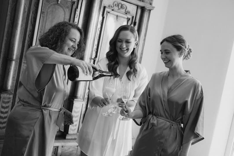 Three women in robes celebrate with champagne, laughing together in a black and white bridal preparation photo.