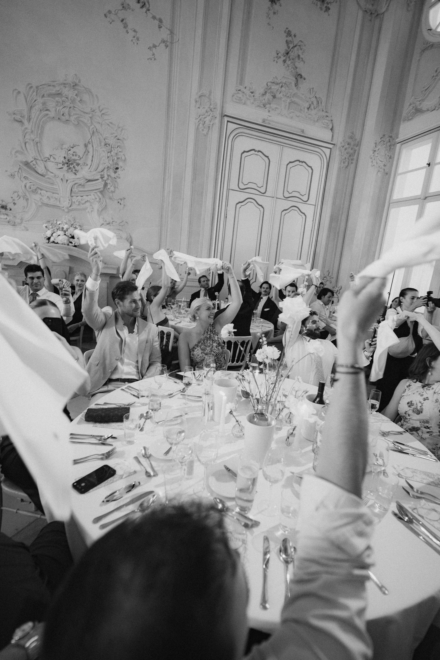 Black and white photo of wedding guests waving napkins in elegant ballroom during celebratory reception dinner.