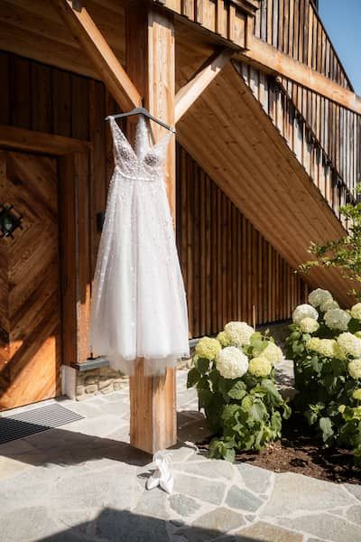Das Brautkleid hängt vor einer Holzhütte. Die Brautschuhe stehen davor.
