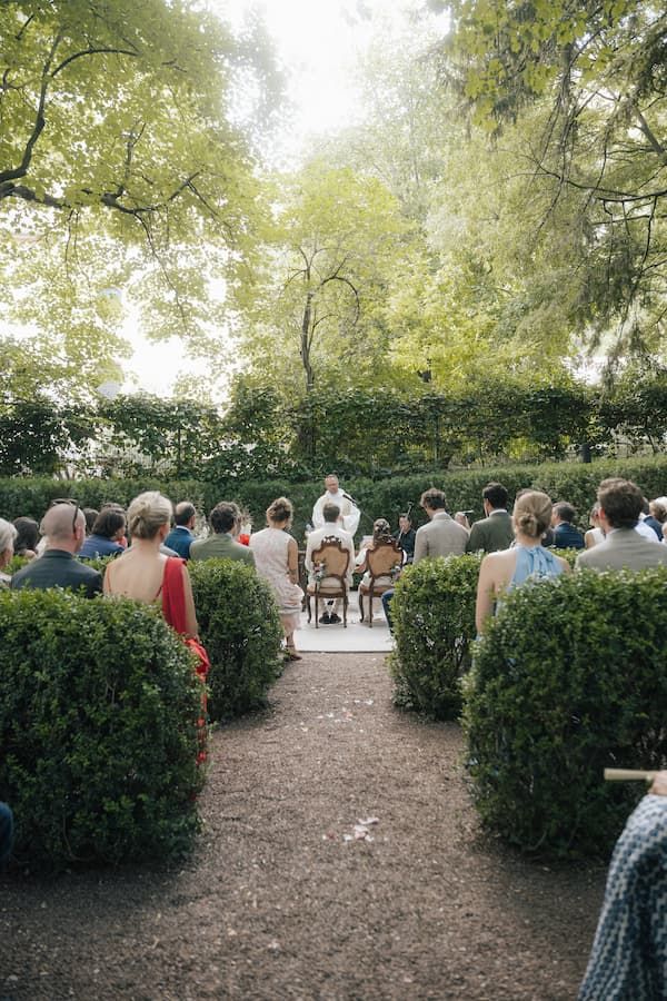 Outdoor wedding ceremony in a lush garden with guests seated along a hedge-lined aisle facing the officiant.