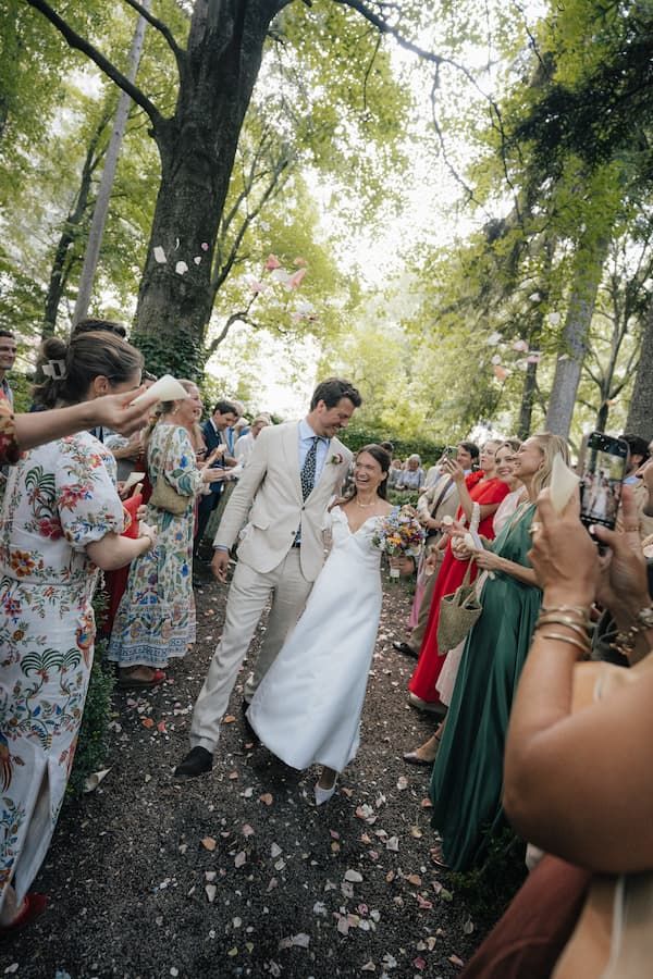 Bride and groom walk through a confetti-lined outdoor aisle surrounded by cheering guests at a garden wedding.