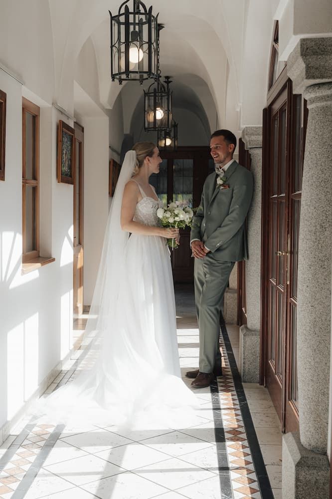 Bride and groom share a joyful moment in a sunlit corridor, bride holding a white bouquet in a flowing wedding dress.