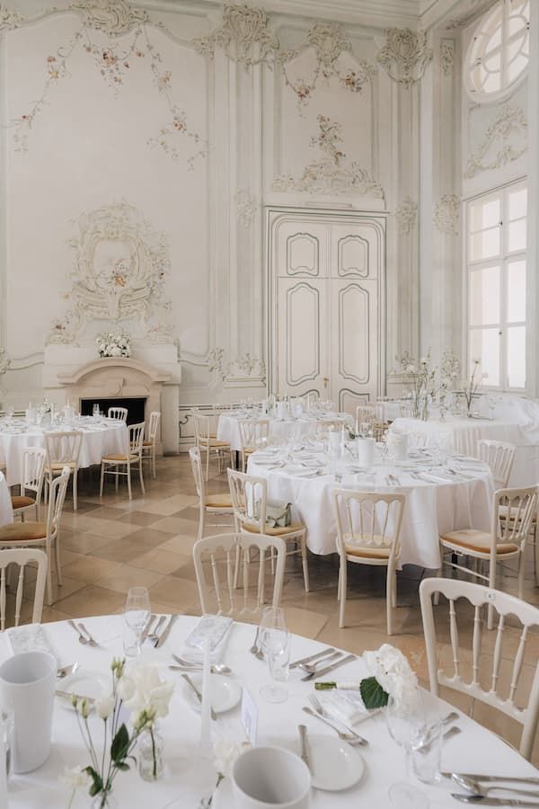 Elegant white wedding reception hall with ornate walls, round tables, and gold chairs.
