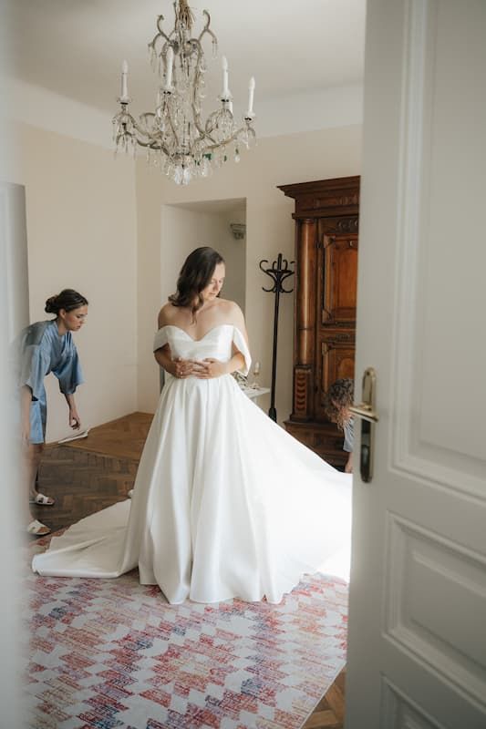 Bride in off-shoulder white ball gown stands in elegant room with chandelier while bridesmaid adjusts her train.