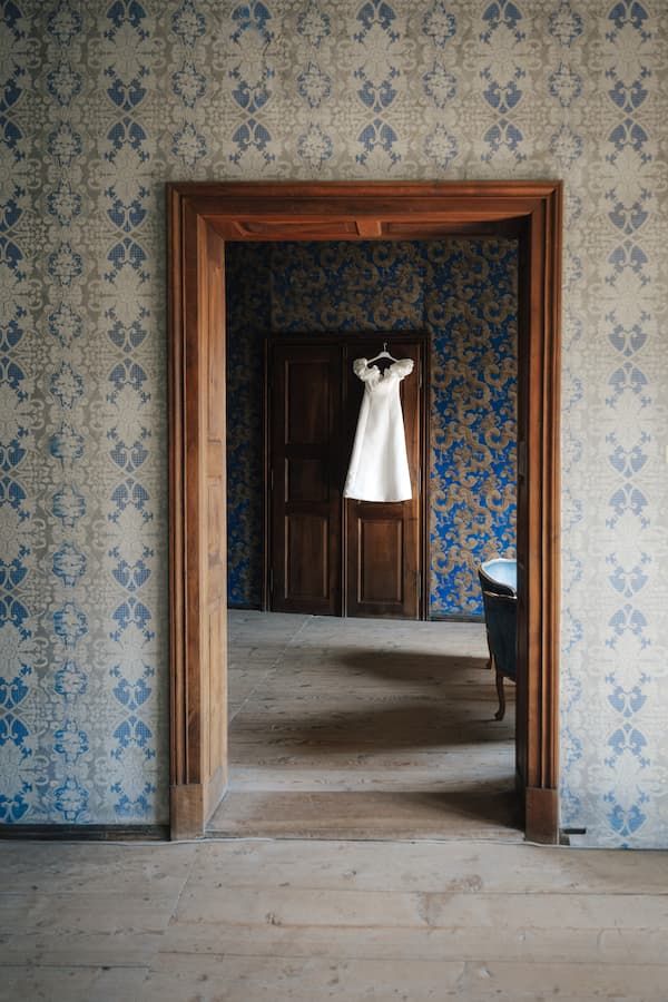 White wedding dress hanging in doorway of vintage room with blue damask wallpaper and hardwood floors.