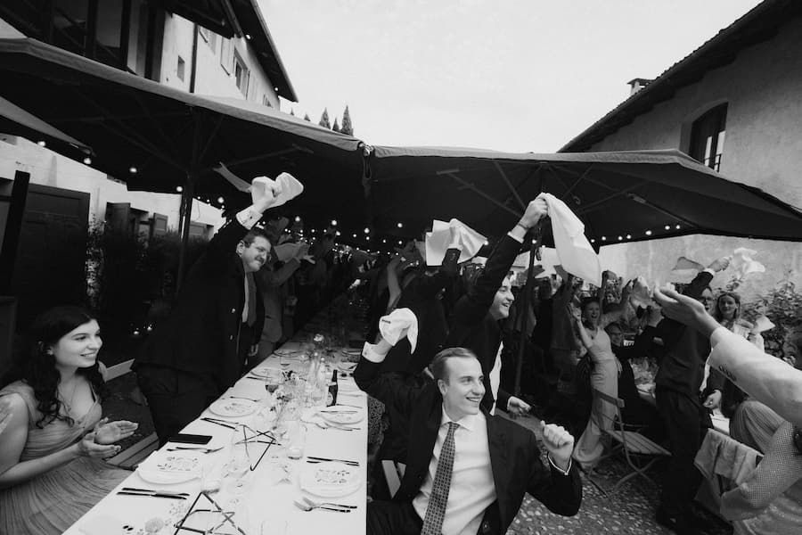 Black and white photo of wedding guests cheering and waving napkins at an outdoor reception dinner table.