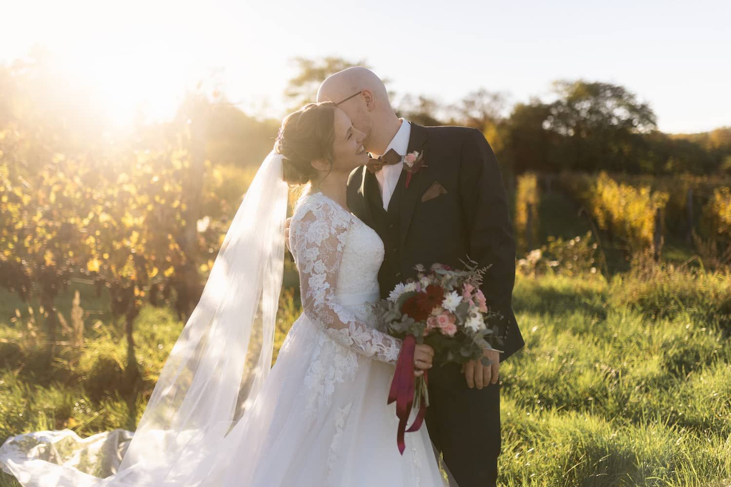 Katrin und Christoph im Brautkleid und Anzug im Weingarten, Christoph flüstert seiner Frau etwas ins Ohr.