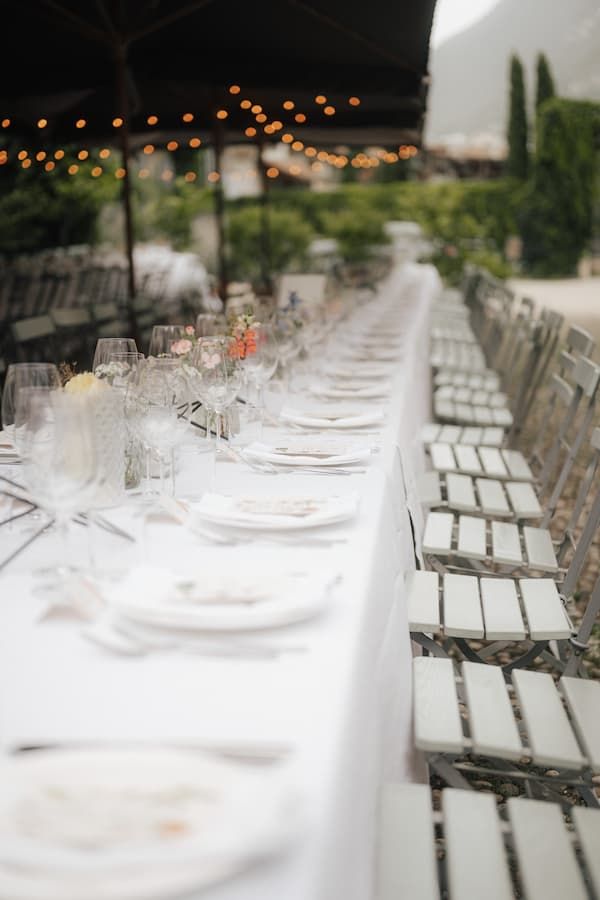 Elegant outdoor wedding reception with long white-draped table, crystal glasses, and string lights overhead.