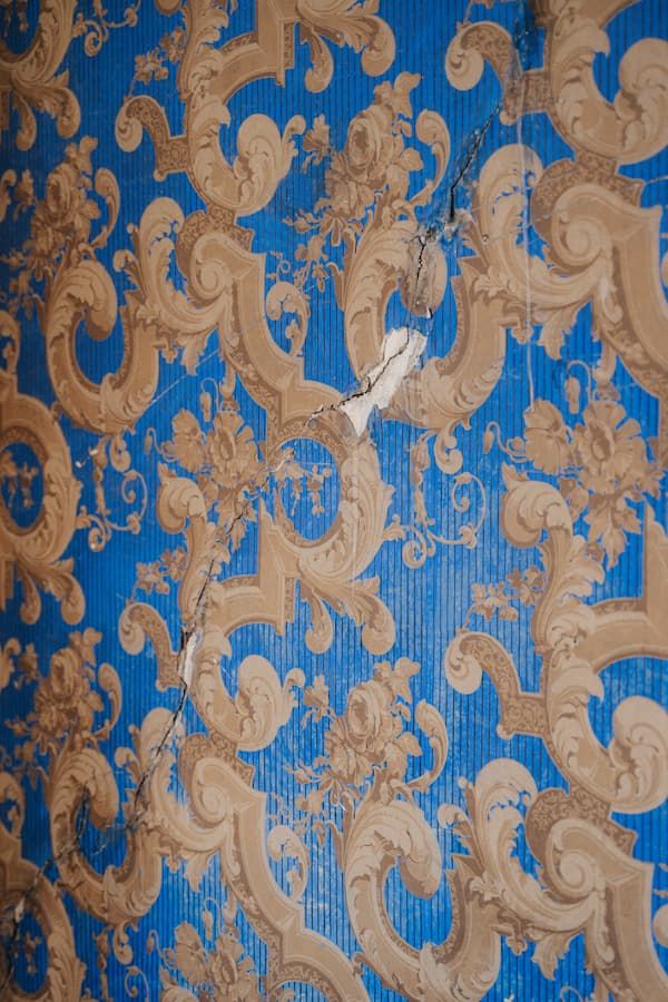 Vintage blue and gold baroque damask wallpaper with ornate scrollwork pattern showing age-related cracking and peeling.