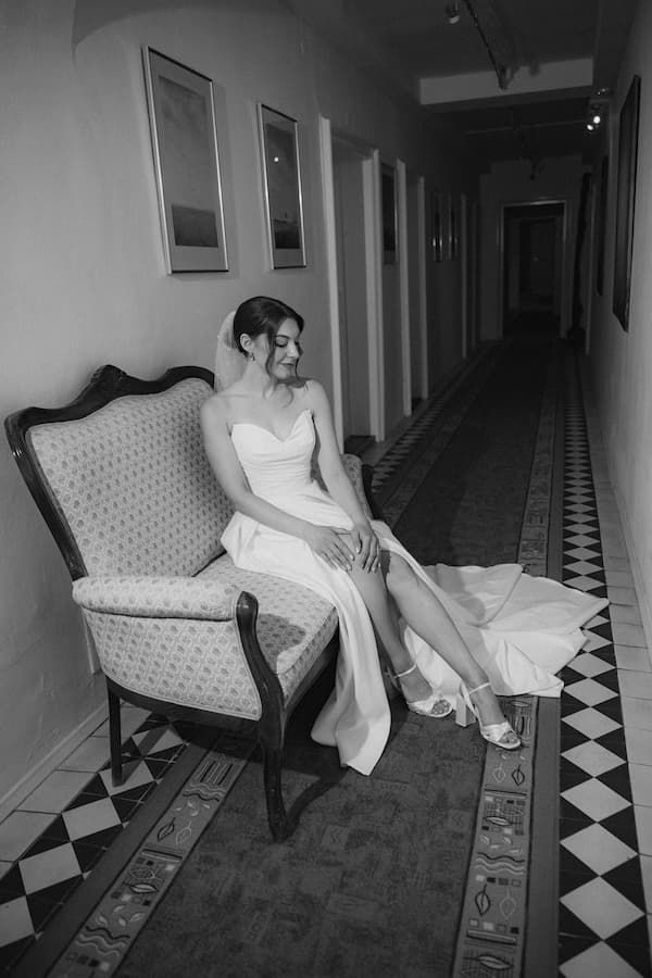 Bride in white gown sits gracefully on vintage chair in elegant hotel hallway, black and white wedding portrait.