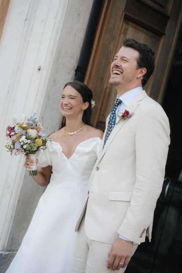 Joyful bride in white off-shoulder gown holding wildflower bouquet stands with laughing groom in cream suit at wedding.