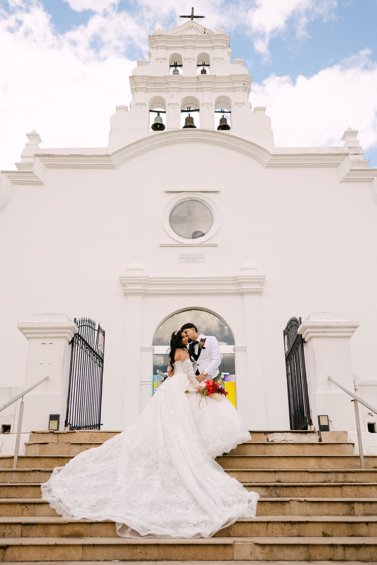 A bride in a flowing white gown poses on stone steps in front of a Spanish colonial style church with bell towers.