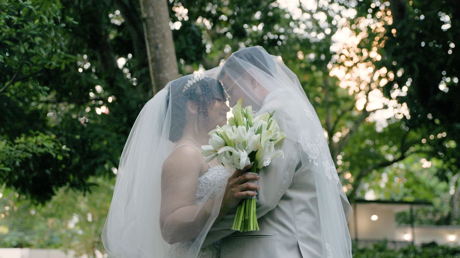 A couple shares an intimate moment under a veil with a bouquet of white flowers.