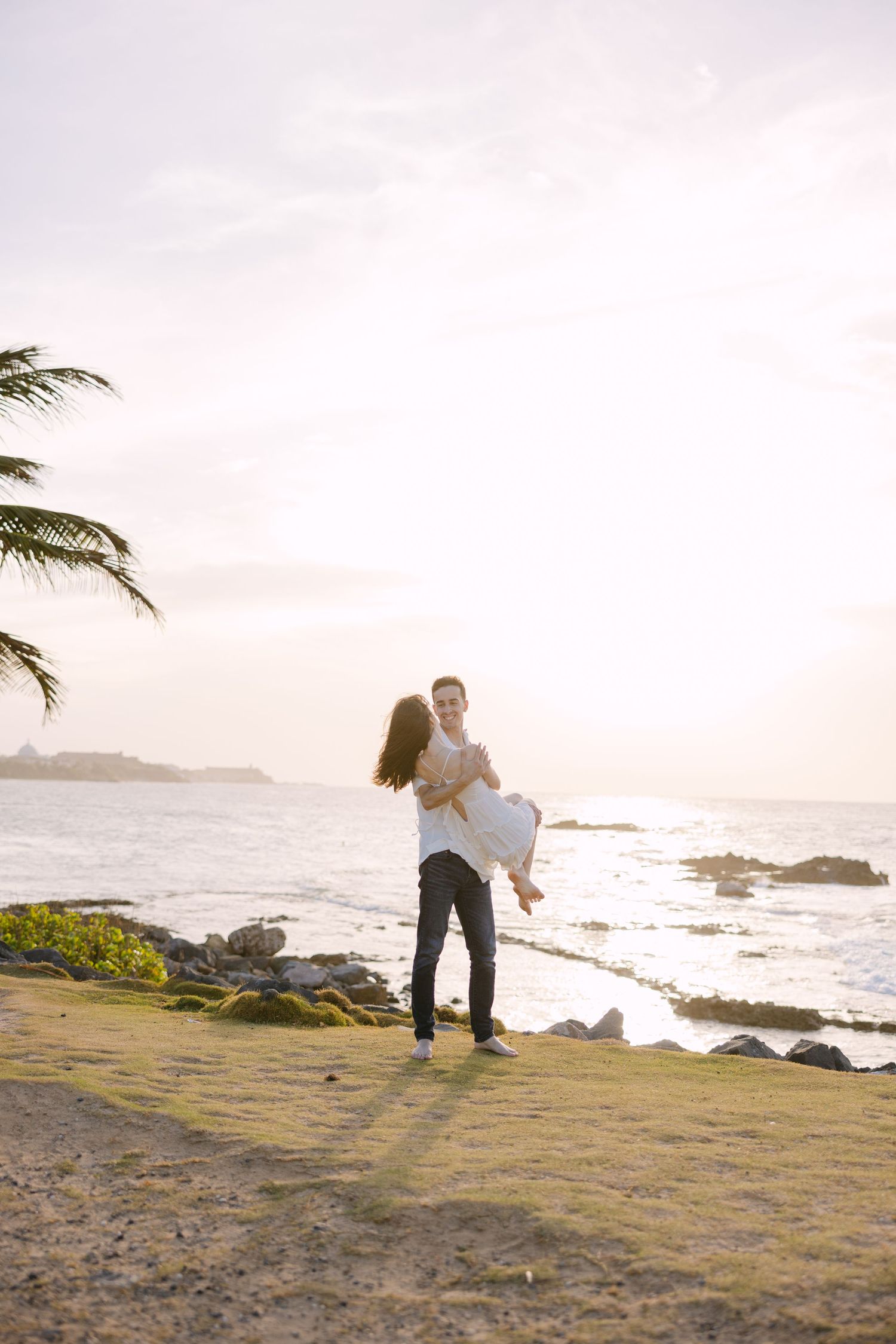 Silhouetted couple embraces on coastal cliff with palm tree and ocean waves at golden hour.